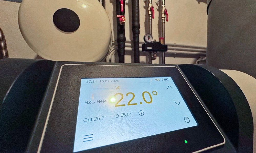 Bedienpanel einer M-TEC-Wärmepumpe mit 22,0 °C Anzeige; dahinter Heizungsrohre, Ventile und Ausdehnungsgefäß im Technikraum.